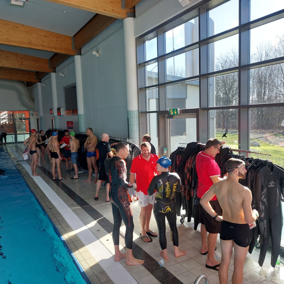 Eine lichtdurchflutete Schwimmhalle: mehrere Menschen stehen am Beckenrad neben Neoprenständern und werden beraten