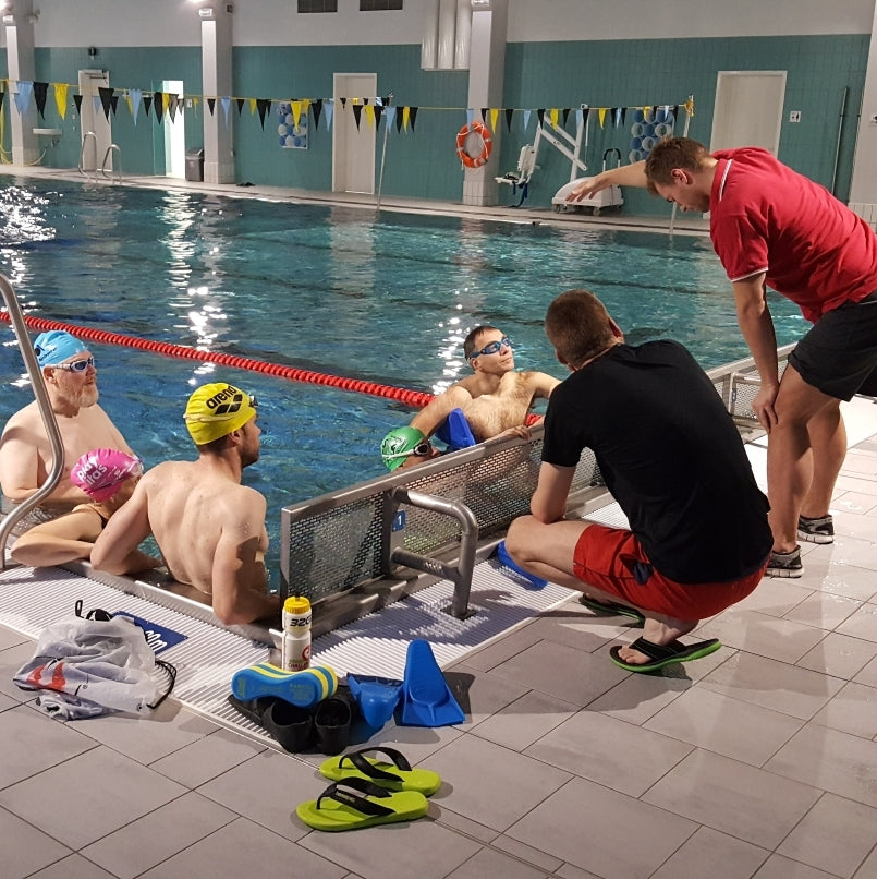 Zwei Schmimmtrainer demonstrieren am Beckenrand den Kraul-Armzug, die Schwimmer im Wasser schauen zu